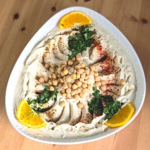 Frisch zubereiteter Hummus mit Kichererbsen, Petersilie, Paprika und Orangenscheiben – serviert auf einem weißen Teller.